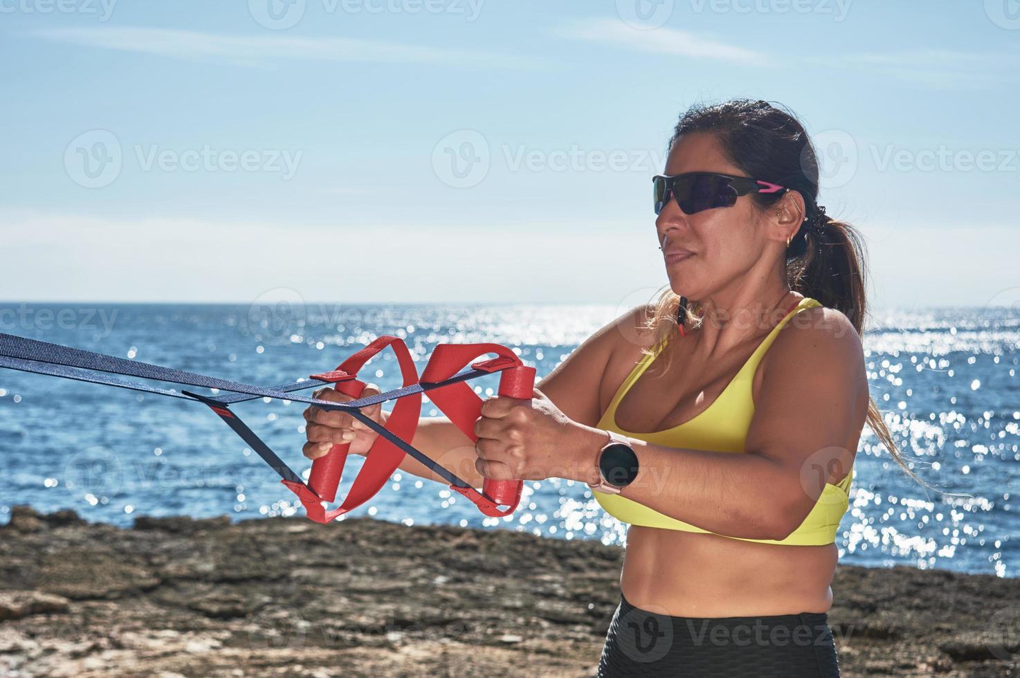 Latin woman, middle-aged, looking at app, smart watch, resting after gym session, wearing yellow top, black leggings, dumbbell, burning calories, staying fit, outdoors by the sea, photo