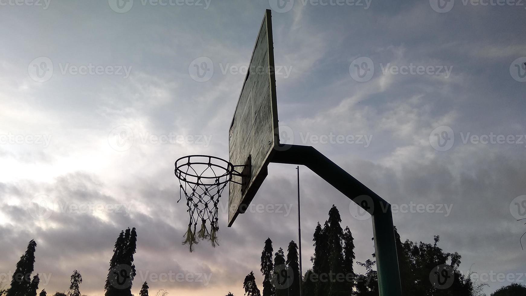 vista lateral del antiguo aro de baloncesto verde tenue y red rota con un fondo oscuro del cielo matutino en el campo deportivo público. foto