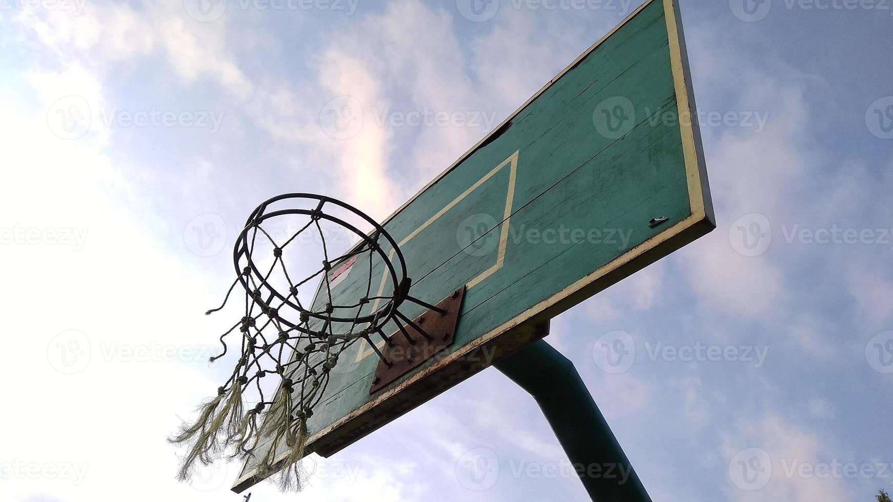 Vista frontal en ángulo bajo del viejo aro de baloncesto verde tenue y red rota con un fondo azul del cielo matutino en el campo deportivo público. foto