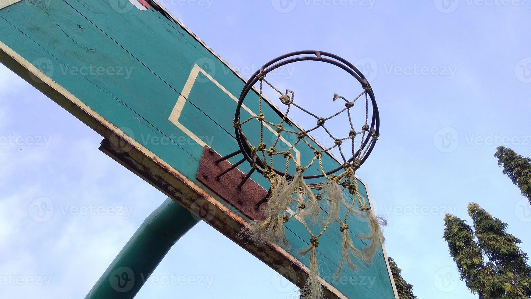 Front-low angle view of dim green old basketball hoop and broken net with a blue background of morning sky in the public sport field. photo