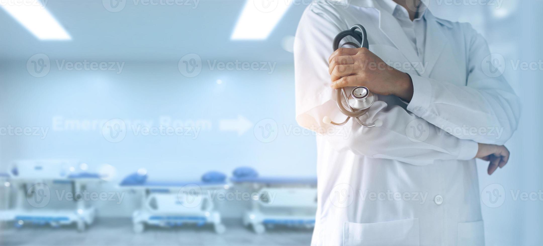 médico con estetoscopio en la mano, parado con confianza frente a la sala de emergencias, camas de pacientes detrás de él, antecedentes hospitalarios foto