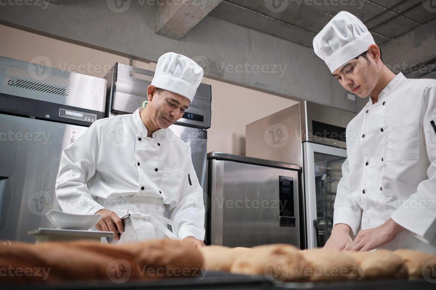 dos chefs asiáticos profesionales con uniformes y delantales de cocinero blanco están amasando masa de pastelería y huevos, preparando pan y comida fresca de panadería, horneando en el horno en la cocina de acero inoxidable del restaurante. foto