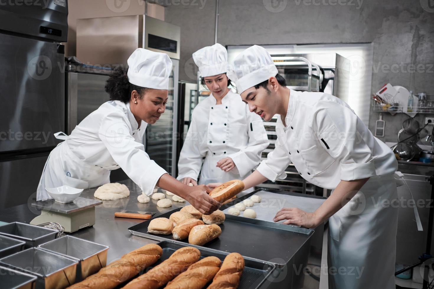 Professional gourmet team, three young chefs in white cook uniforms and aprons knead pastry dough and eggs, prepare bread, and fresh bakery food, baking in oven at stainless steel restaurant kitchen. photo