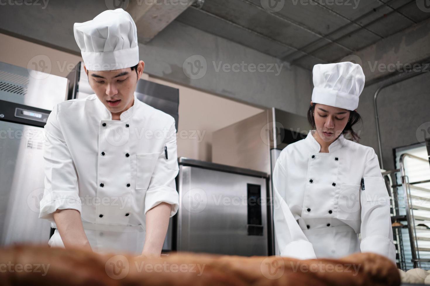 Two professional Asian chefs in white cook uniforms and aprons knead pastry dough and eggs, prepare bread, cookies, and fresh bakery food, baking in an oven in a restaurant stainless steel kitchen. photo
