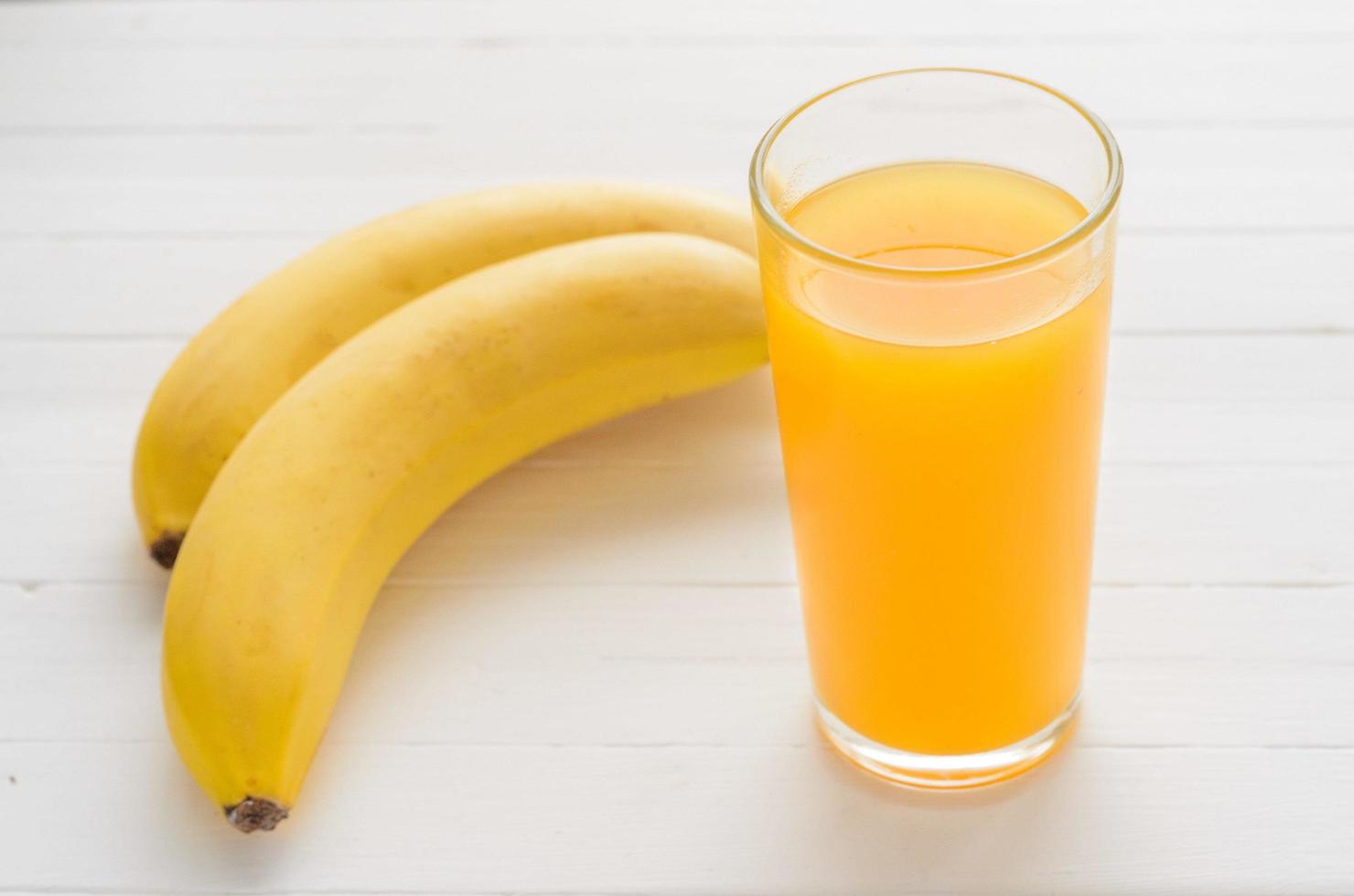 vaso de jugo de frutas múltiples y dos plátanos en una mesa de madera blanca. foto