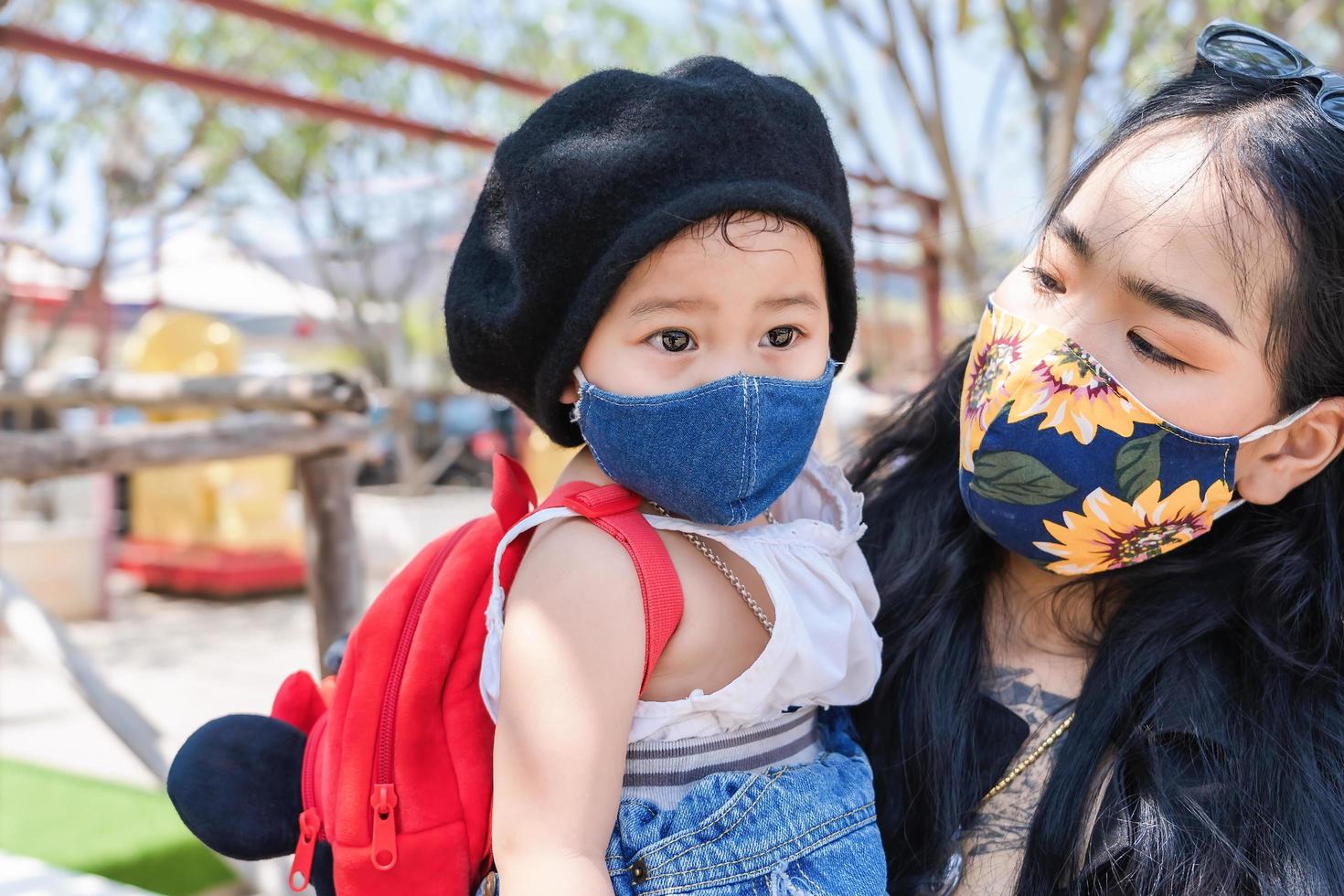 Little child girl and Asian mother wearing medical protective mask and holding her daughter in a crowded public place in park or tourist. photo