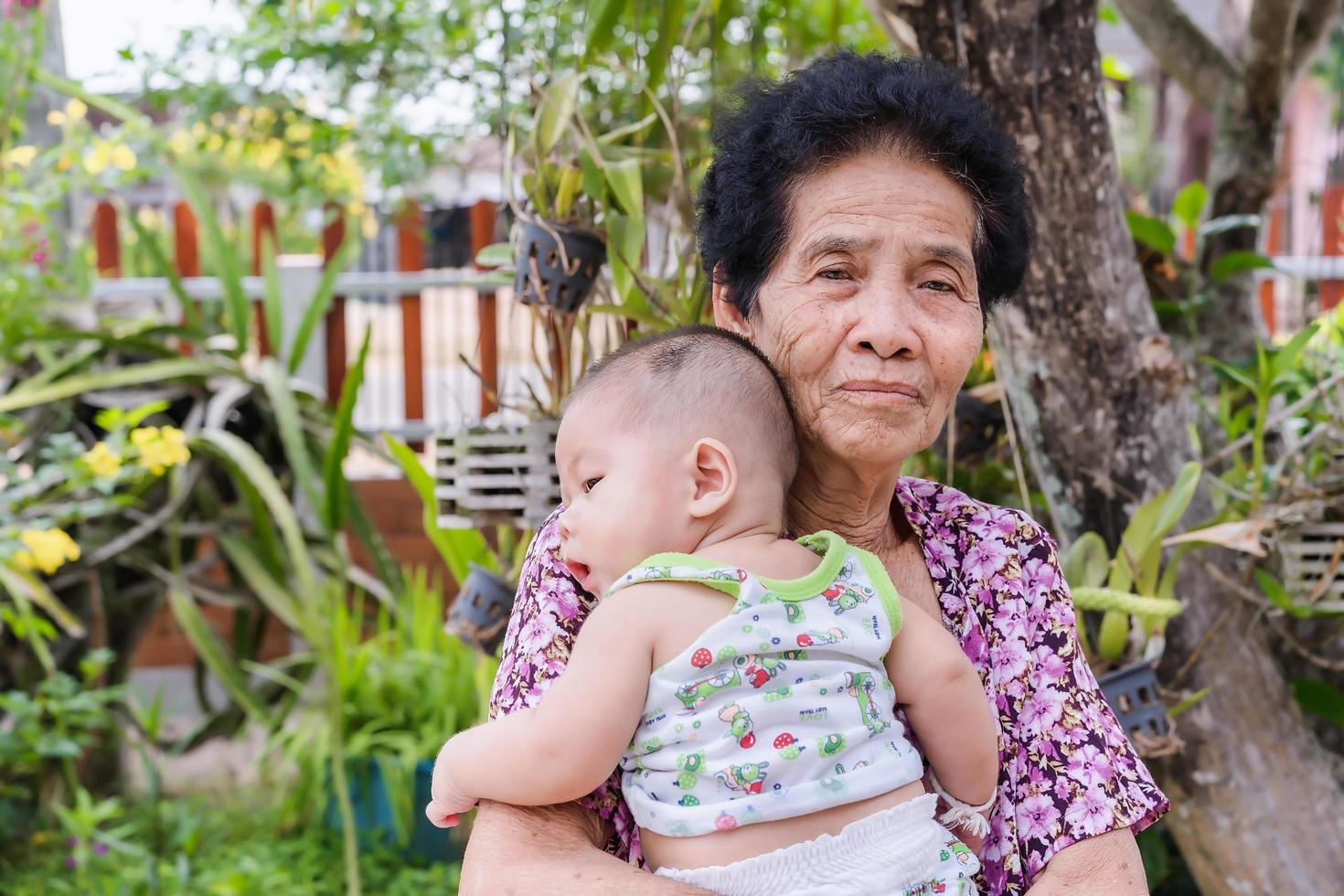 Happy senior woman holding adorable baby boy in spring flowery garden. Grandmother holding newborn baby grandson looking to camera outdoors photo