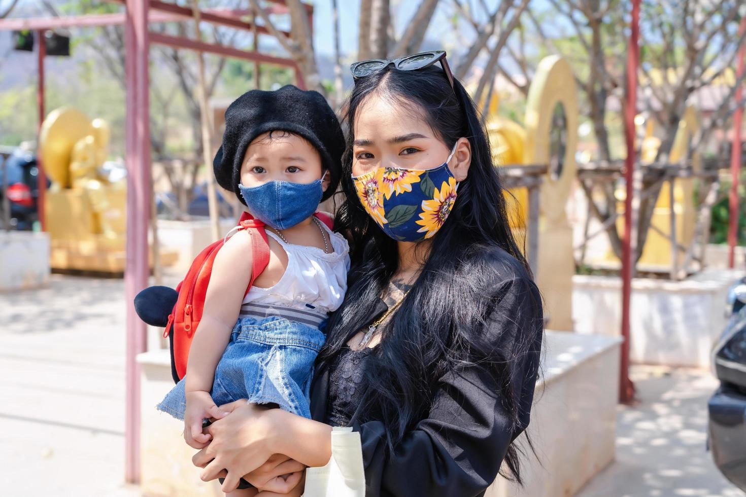 Little child girl and Asian mother wearing medical protective mask and holding her daughter in a crowded public place in park or tourist. photo