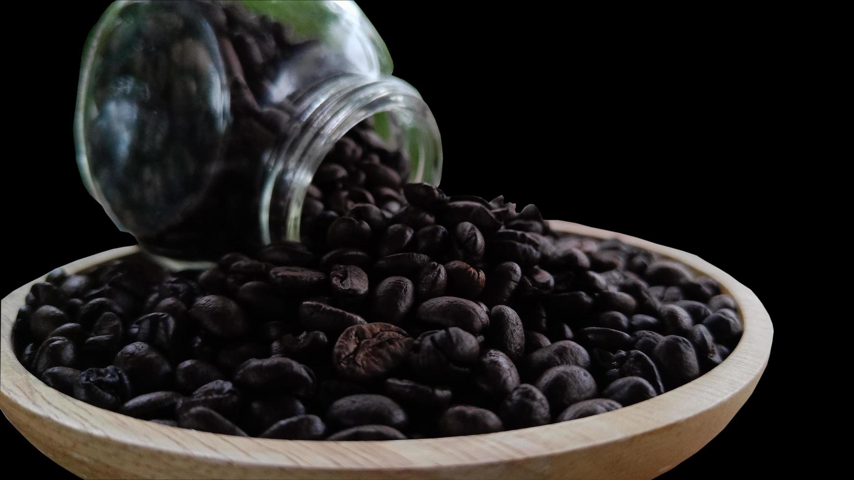 close up side view Coffee beans from a glass bottle on a wooden plate on a black background photo