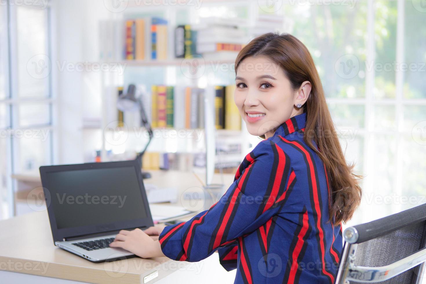 una hermosa mujer asiática profesional que usa una camisa de manga larga se sienta en una silla para trabajar sonriendo alegremente en la oficina, está mirando la cámara que tiene una computadora portátil puesta en la mesa. foto