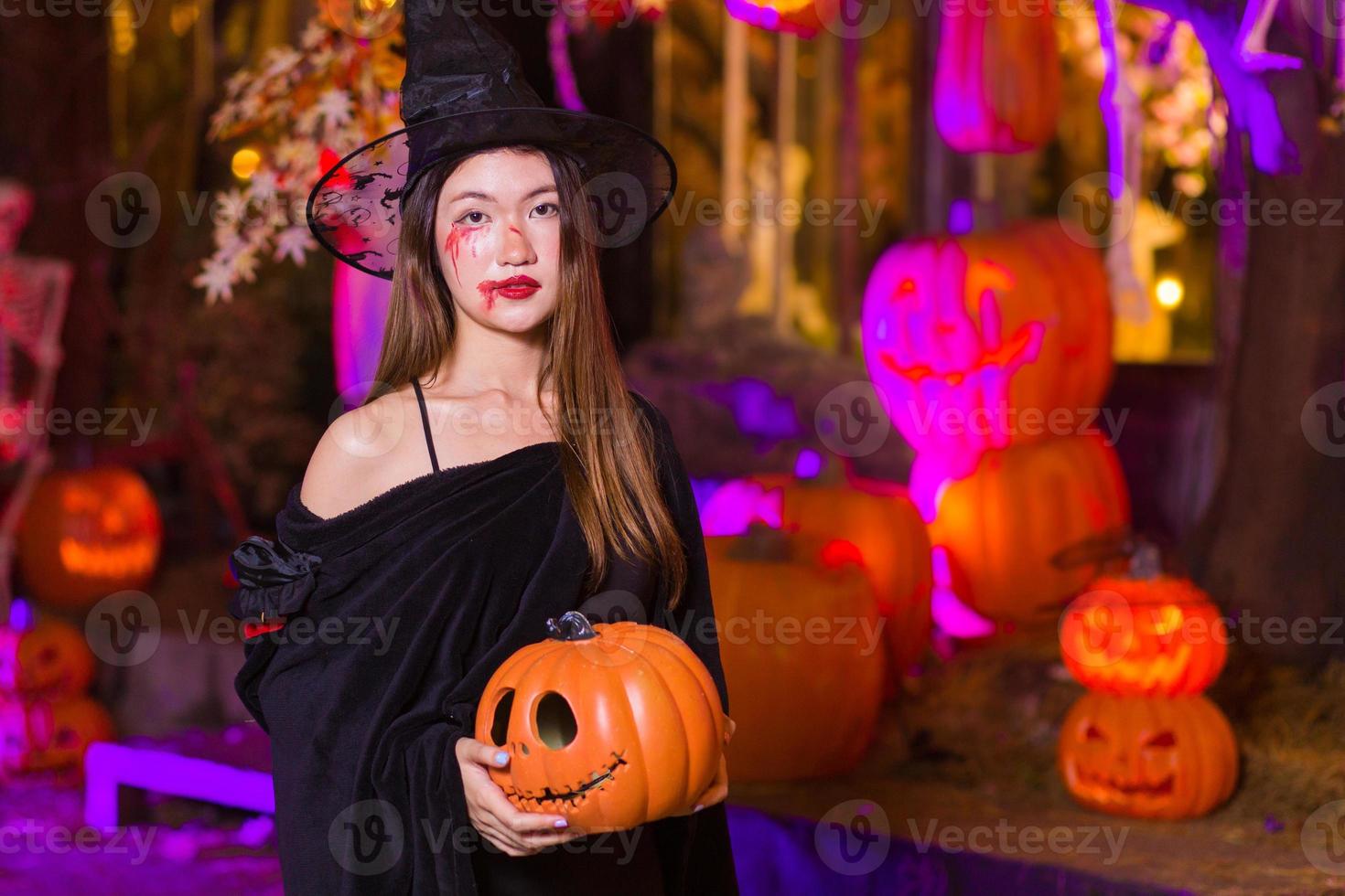 una mujer asiática usa un abrigo negro fantasma y se maquilla como un fantasma de bruja mientras mira la cámara y sostiene una linterna de cabeza de calabaza en sus manos por la noche en el fondo exterior de calabaza en el día de halloween. foto