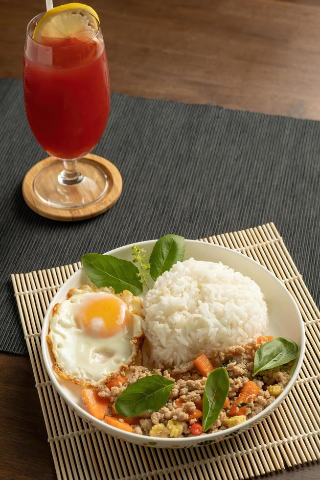Stir-fried Thai basil with minced pork meat, carrots, and baby corns, with fried egg and fresh basils sort in a white dish on bamboo placemat, grey placemat, brown wood table. Copy space photo