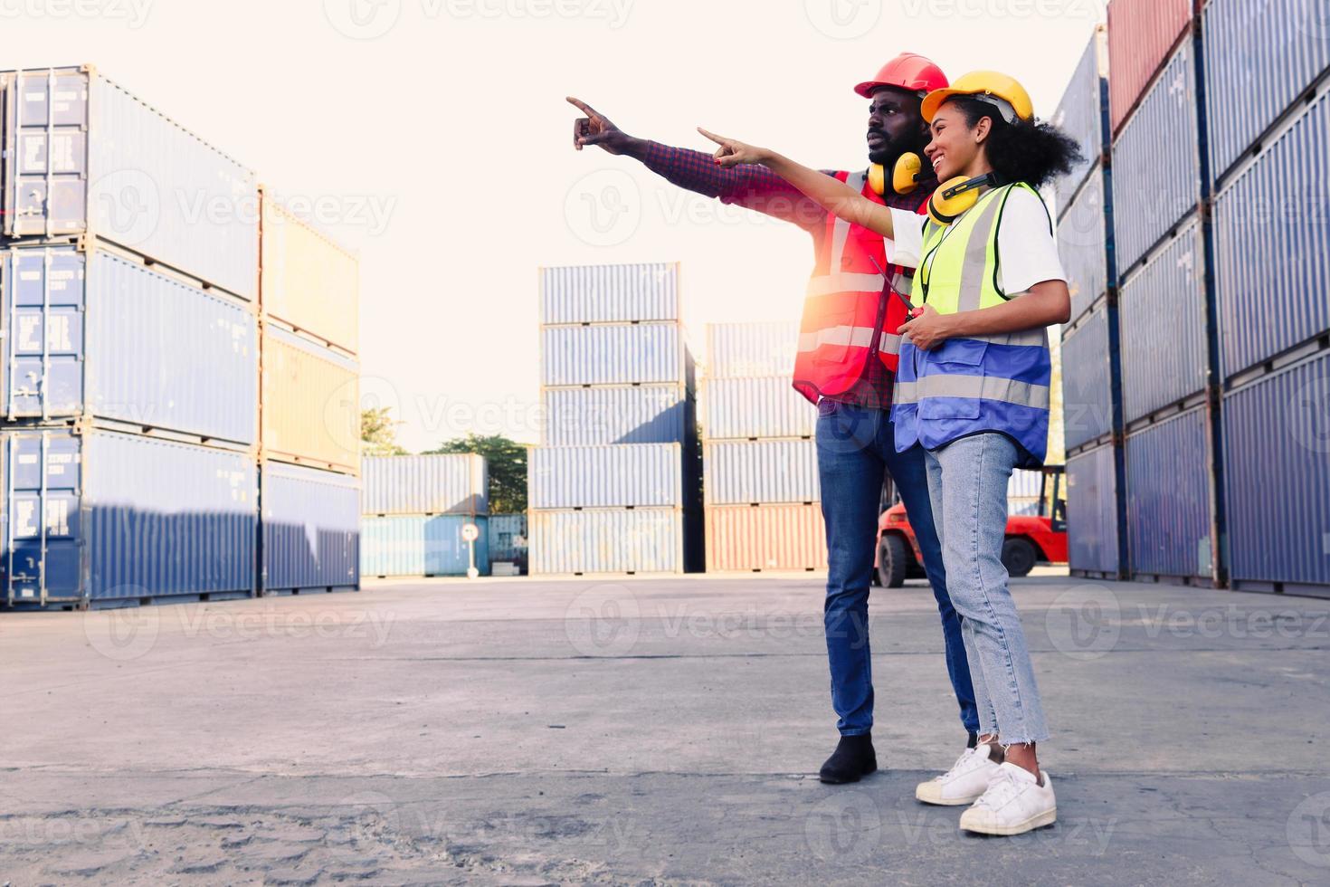 dos felices y sonrientes ingenieros industriales afroamericanos, hombre y mujer, con chaleco de seguridad y casco, señalando hacia afuera, trabajadores que trabajan en el lugar de trabajo del patio de carga de envío logístico. foto