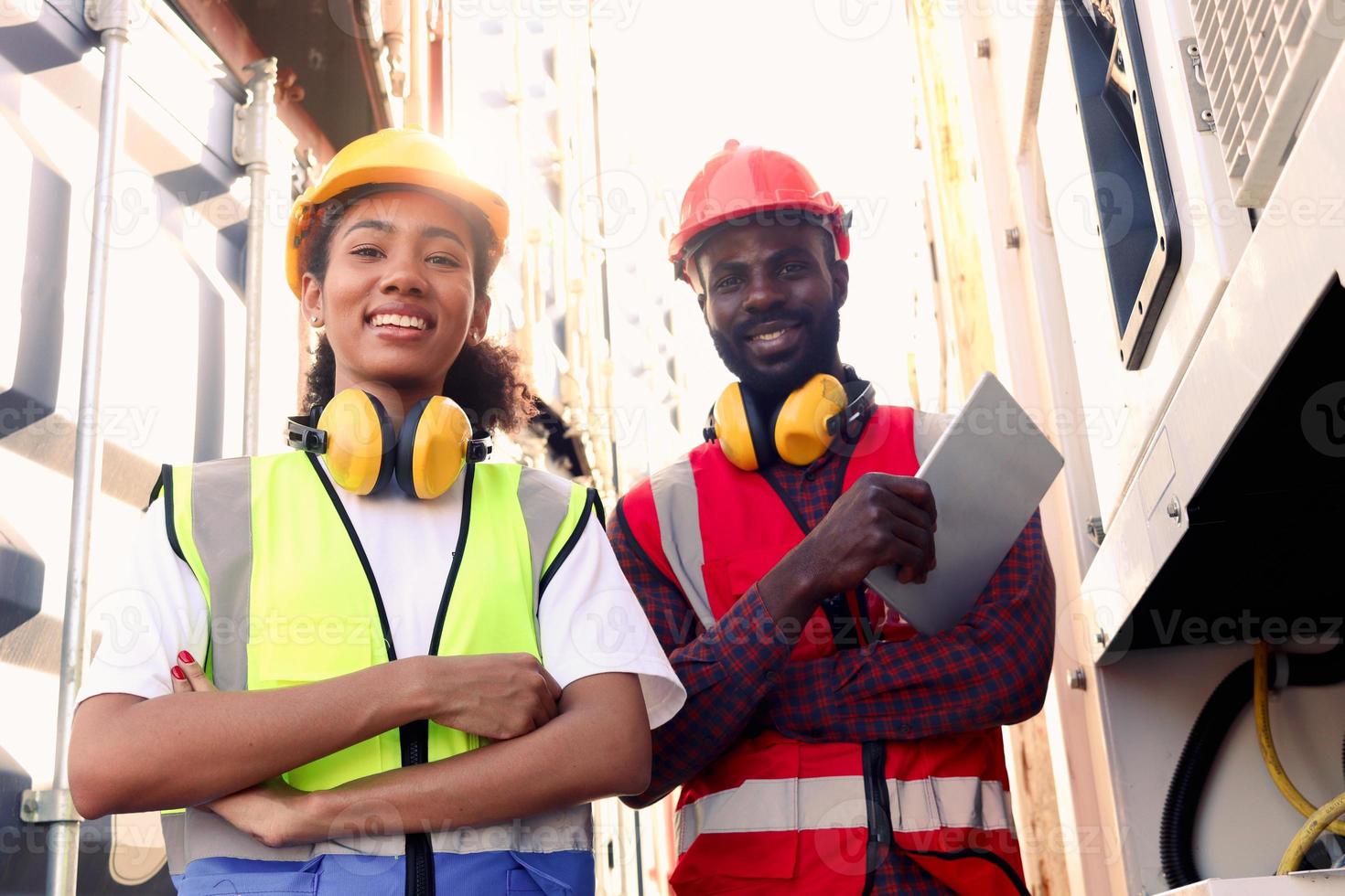 retrato de dos felices y sonrientes trabajadores industriales afroamericanos, un hombre y una mujer, con chaleco de seguridad y casco, de pie con los brazos cruzados en el patio de contenedores de carga logística. foto