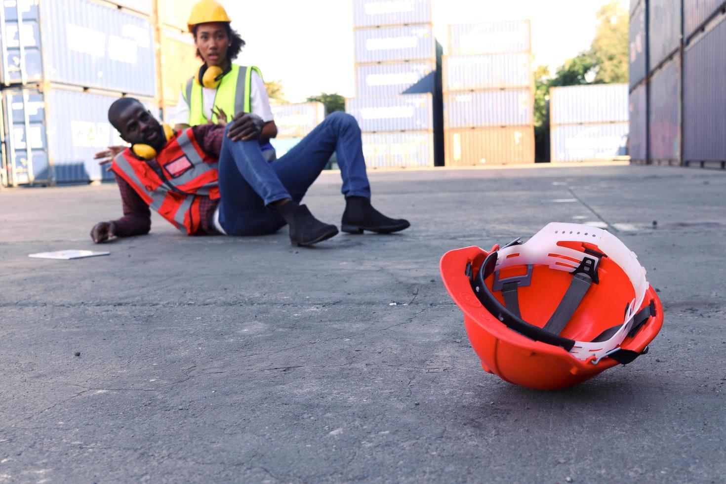 Safety helmet on ground with background of industrial African American engineer worker man getting hurt from accident at workplace, lying on floor at construction site area, colleague try to help him. photo