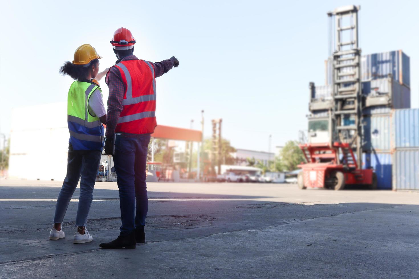 Behind two industrial African American engineer man and woman wearing safety vest and helmet, pointing and looking at container lifting truck, workers working at logistic shipping cargo yard workplace photo