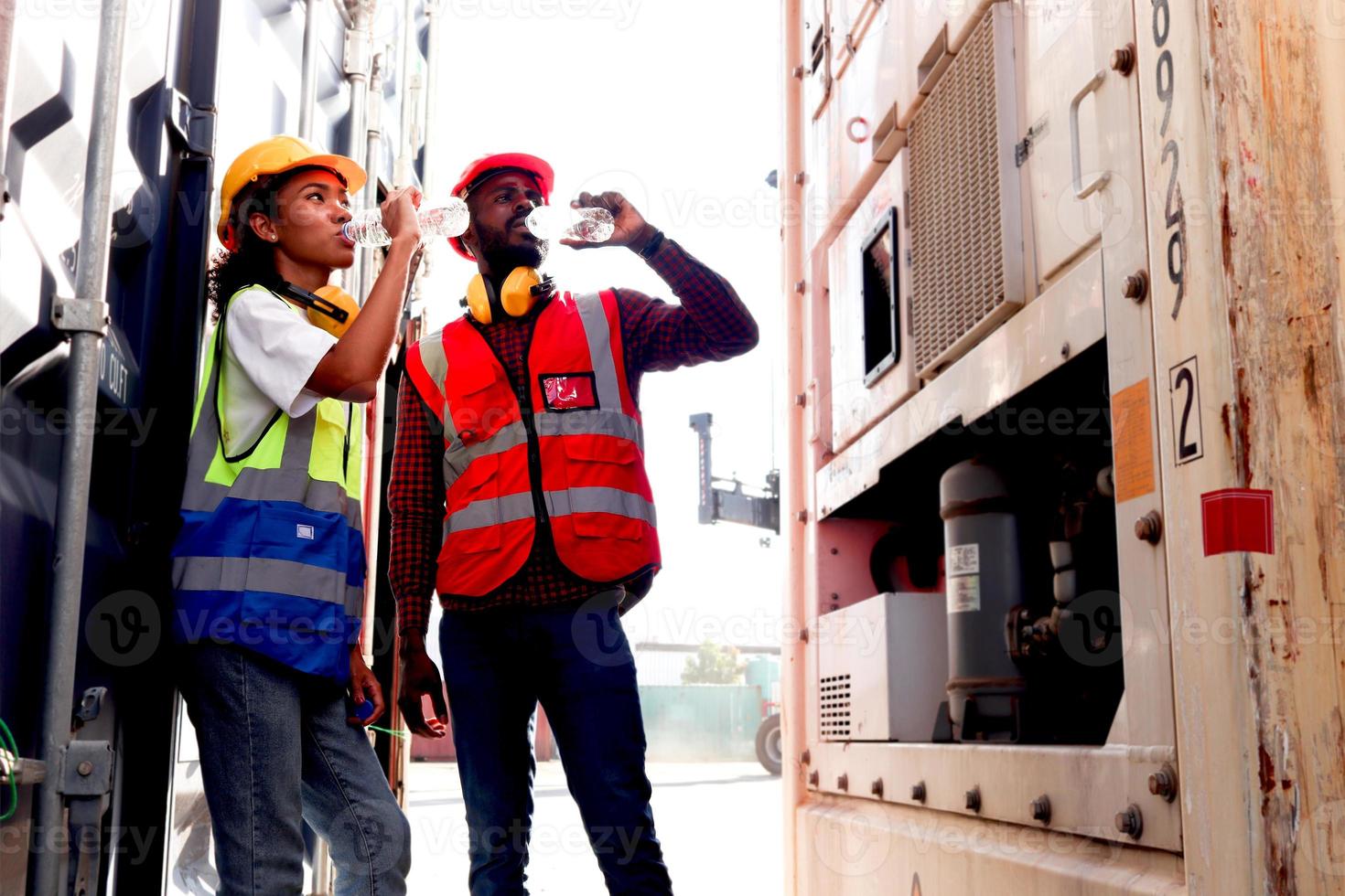 dos ingenieros industriales afroamericanos, un hombre y una mujer que usan chaleco y casco de seguridad, beben agua de una botella durante un descanso después de trabajar duro en el patio de contenedores de carga de envío logístico. foto