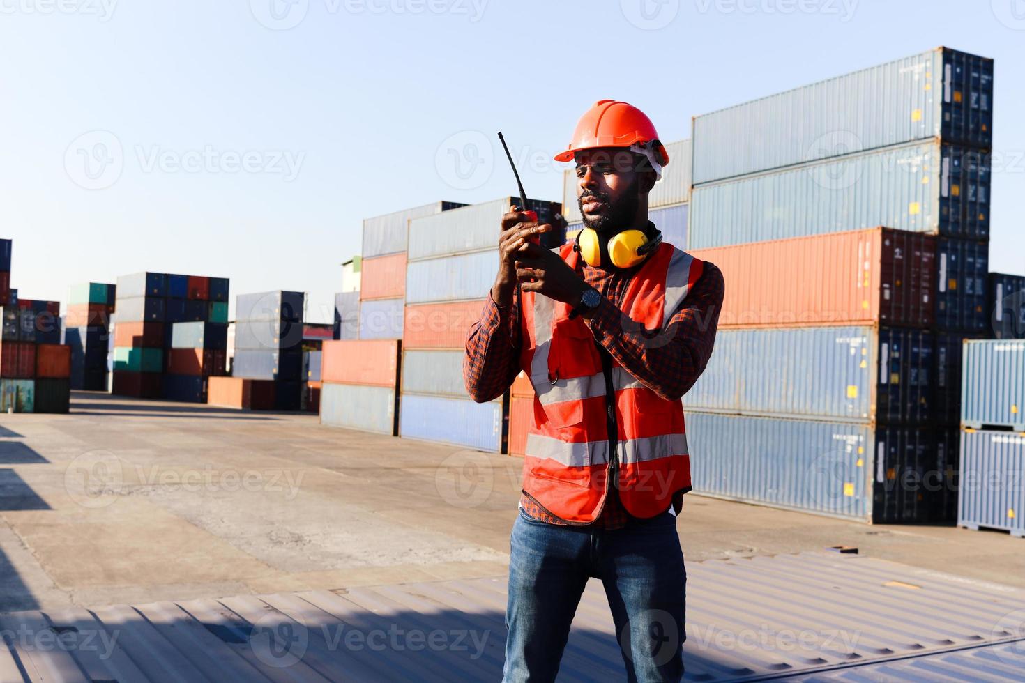 Portrait of African American young engineer worker man wearing safety bright neon red color vest and helmet, talking to a colleague with walkies-talkie at logistic shipping cargo container yard. photo