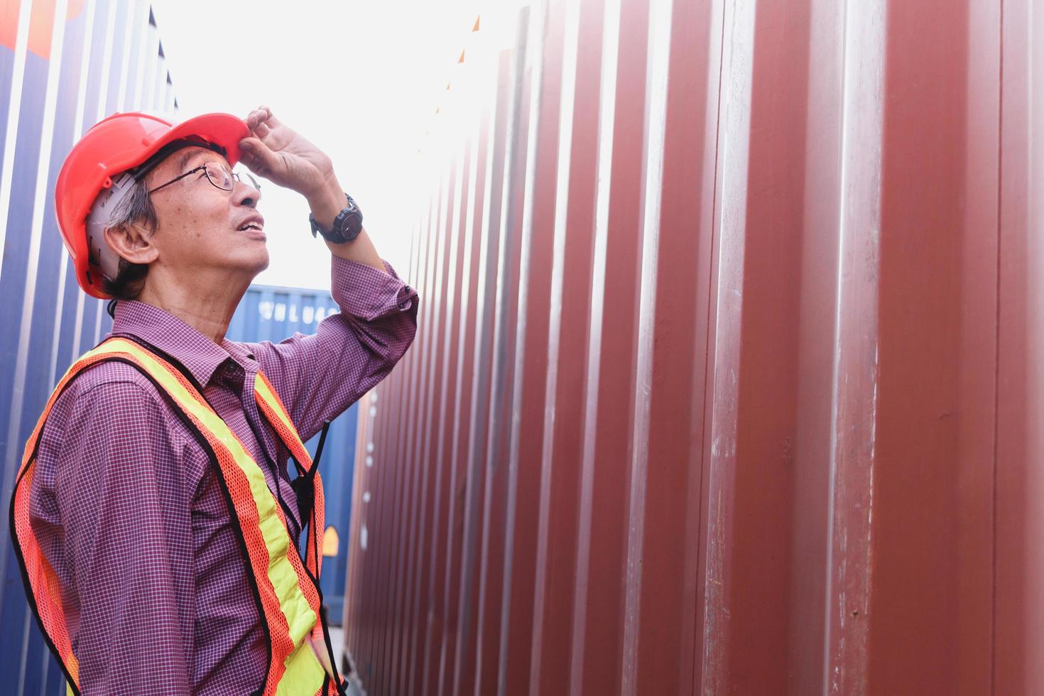 Portrait of senior elderly Asian worker engineer wearing safety vest and helmet, standing between red and blue containers background at logistic shipping cargo containers yard. photo