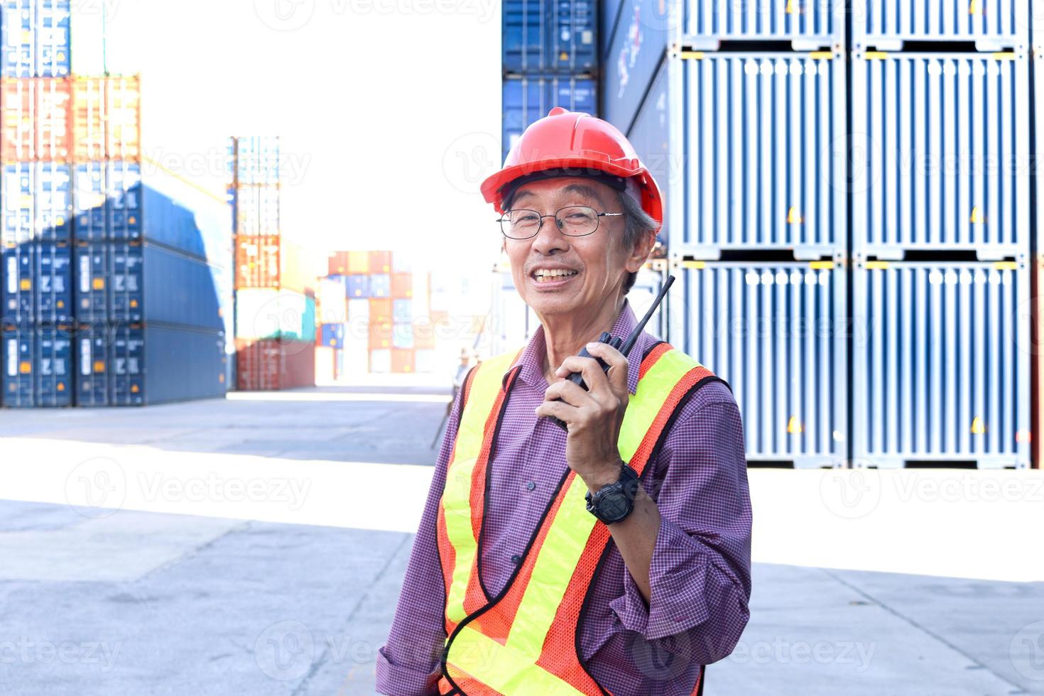 A senior elderly Asian worker engineer wearing safety vest and helmet standing and holding radio walkies talkie at logistic shipping cargo containers yard. elderly people at workplace concept. photo