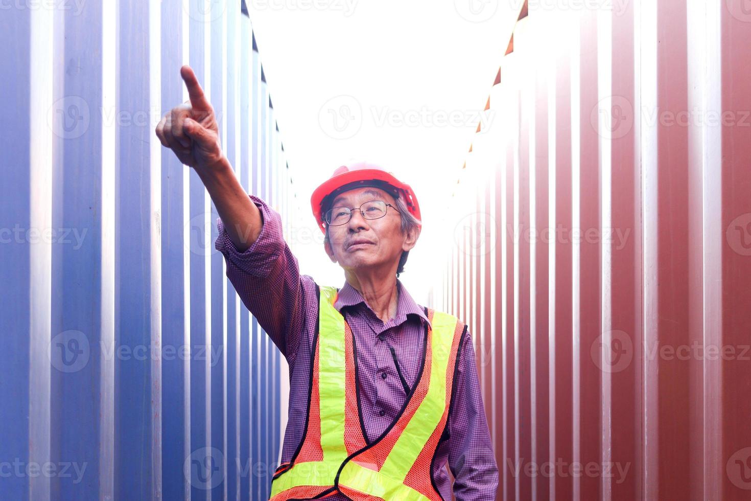 Portrait of senior elderly Asian worker engineer wearing safety vest and helmet, pointing to camera, standing between red and blue containers background at logistic shipping cargo containers yard. photo