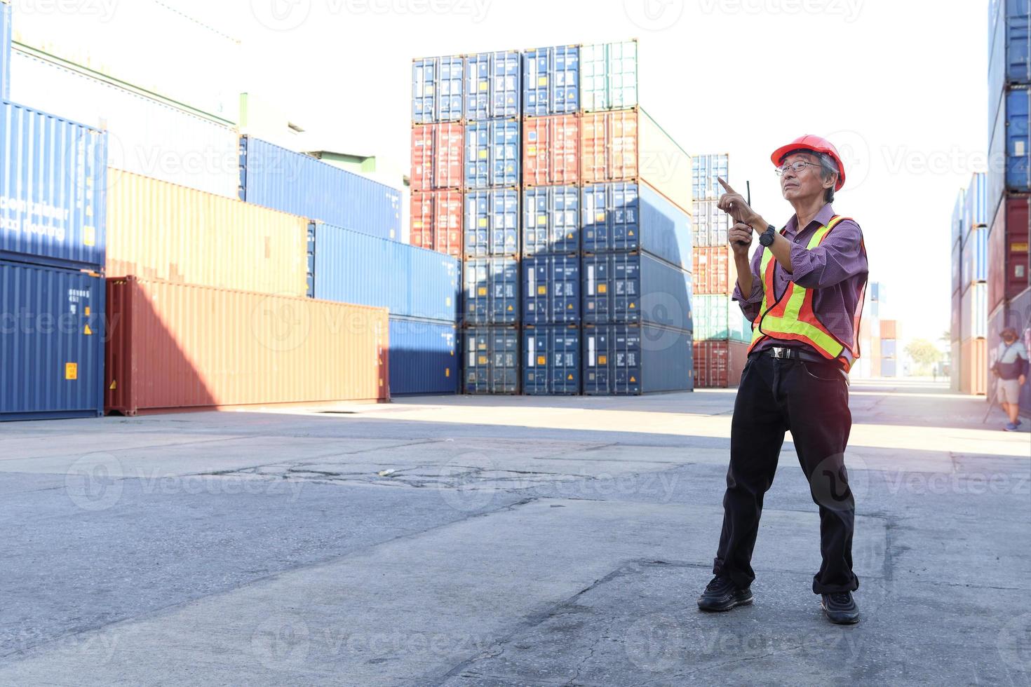 Senior elderly Asian worker engineer wearing safety vest and helmet, holding radio walkies talkie and pointing something at logistic shipping cargo containers yard. elderly people at workplace concept photo