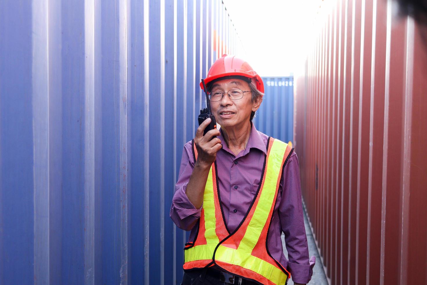 Portrait of senior elderly Asian worker engineer wearing safety vest and helmet, holding radio walkies talkie, standing between red and blue containers at logistic shipping cargo containers yard. photo