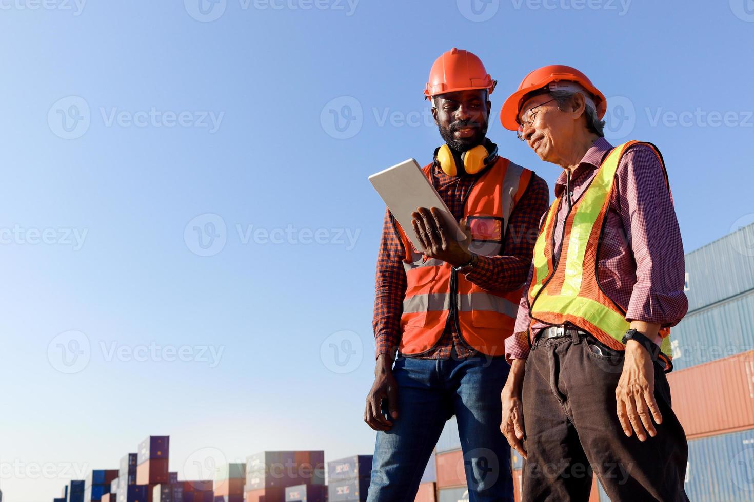 dos trabajadores que usan chaleco de seguridad y casco que usan una tableta digital para discutir en el patio de contenedores de carga de envío logístico. El ingeniero afroamericano y el anciano asiático trabajan juntos. foto