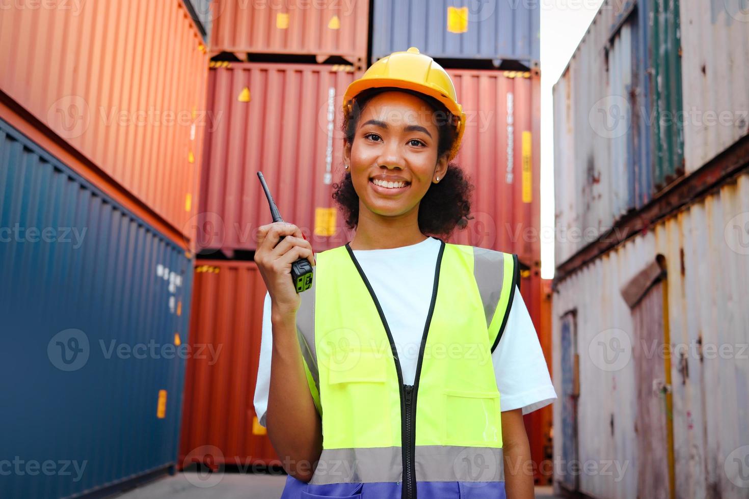 Portrait of happy smiling beautiful African American woman engineer worker with curly hair wearing safety vest and helmet, holding walkies-talkie at logistic shipping cargo container yard. photo
