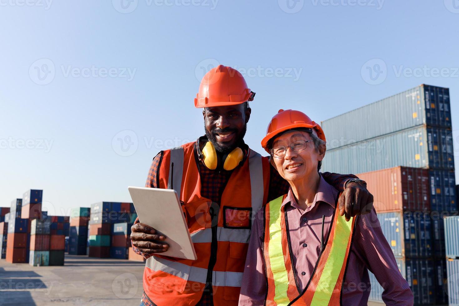 Two workers wear safety helmet work together at logistic shipping cargo containers yard. African American and senior elderly Asian engineer hug each other, respect in differences of race skin color. photo