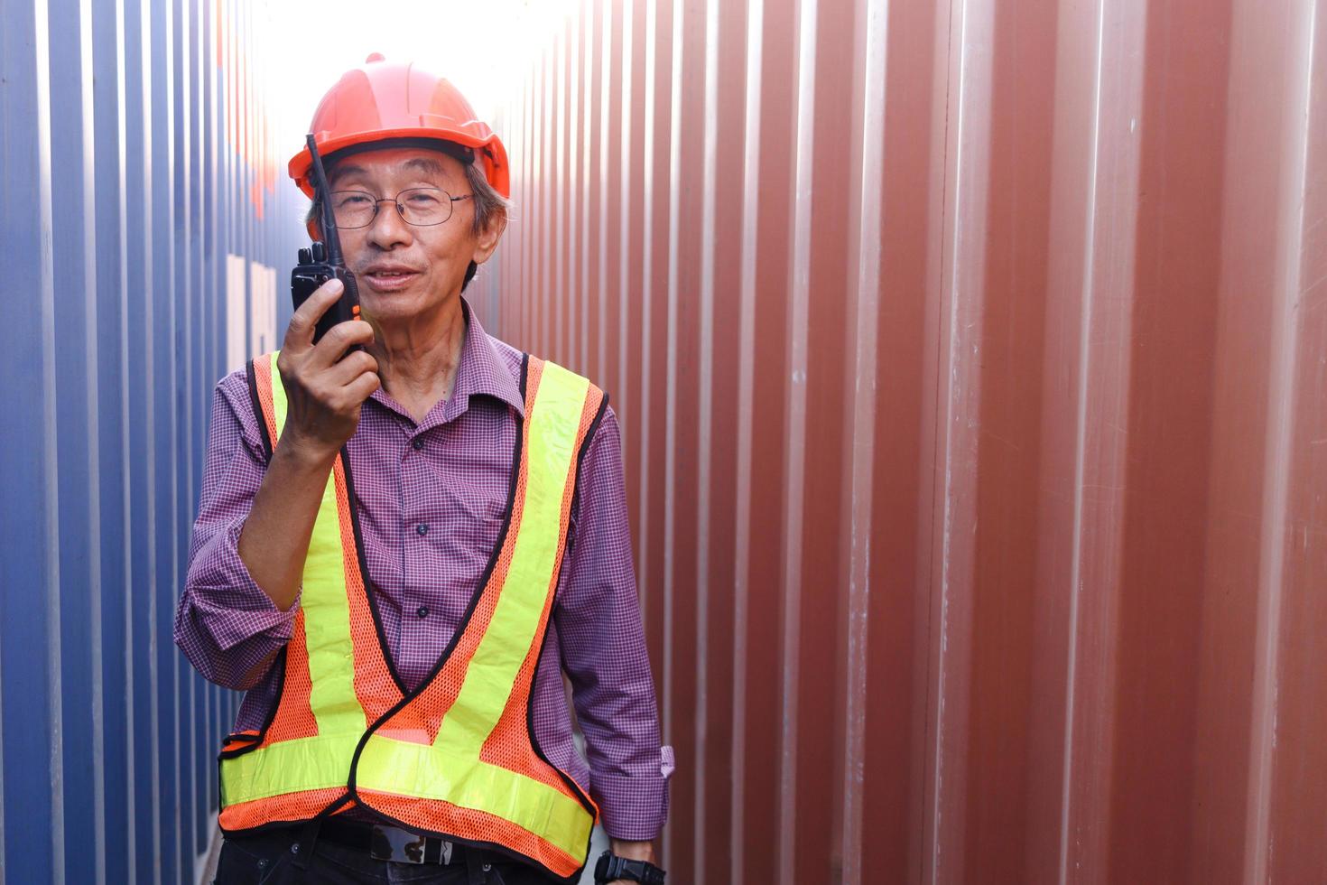 Portrait of senior elderly Asian worker engineer wearing safety vest and helmet, holding radio walkies talkie, standing between red and blue containers at logistic shipping cargo containers yard. photo