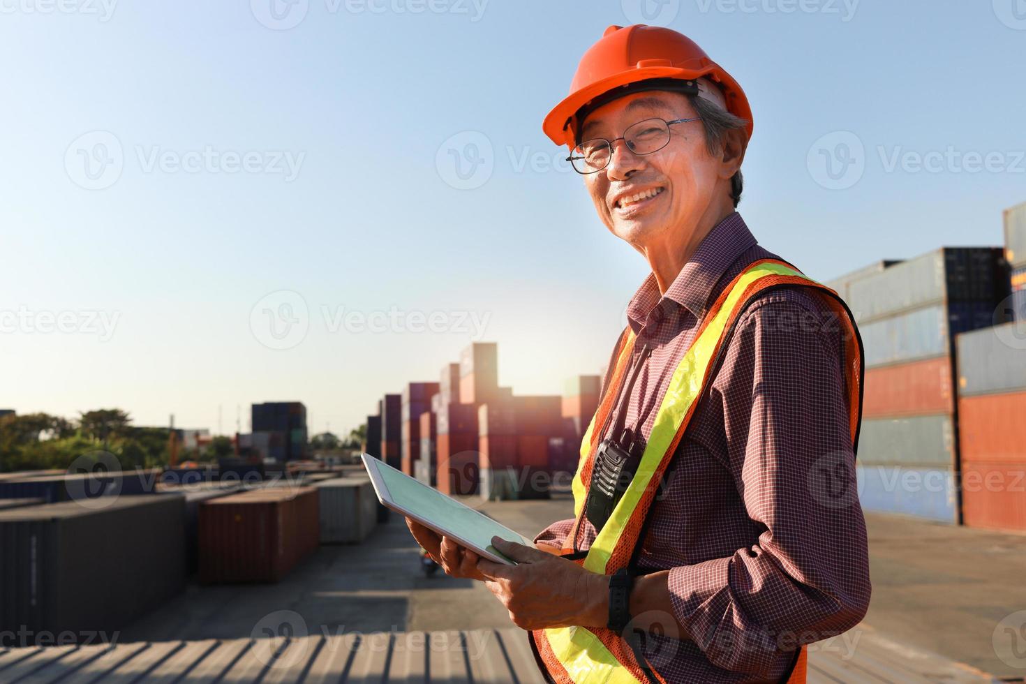 A senior elderly Asian worker engineer wearing safety vest and helmet standing and holding digital tablet at shipping cargo containers yard. elderly people at workplace concept photo