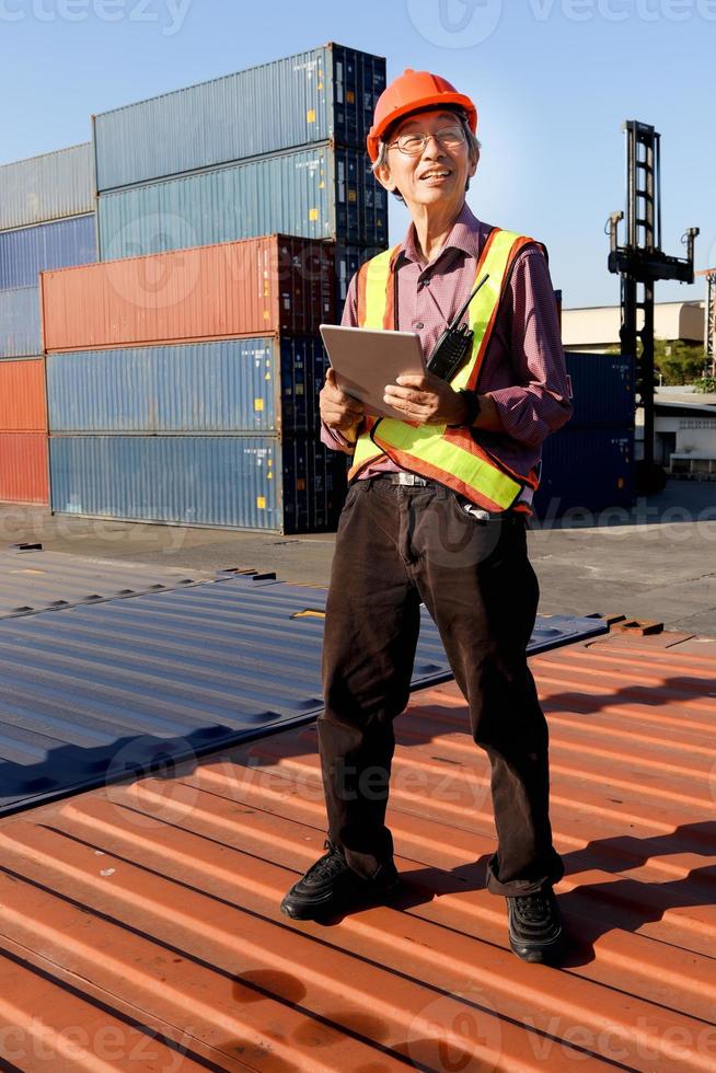 A senior elderly Asian worker engineer wearing safety vest and helmet standing and holding digital tablet at shipping cargo containers yard. elderly people at workplace concept photo