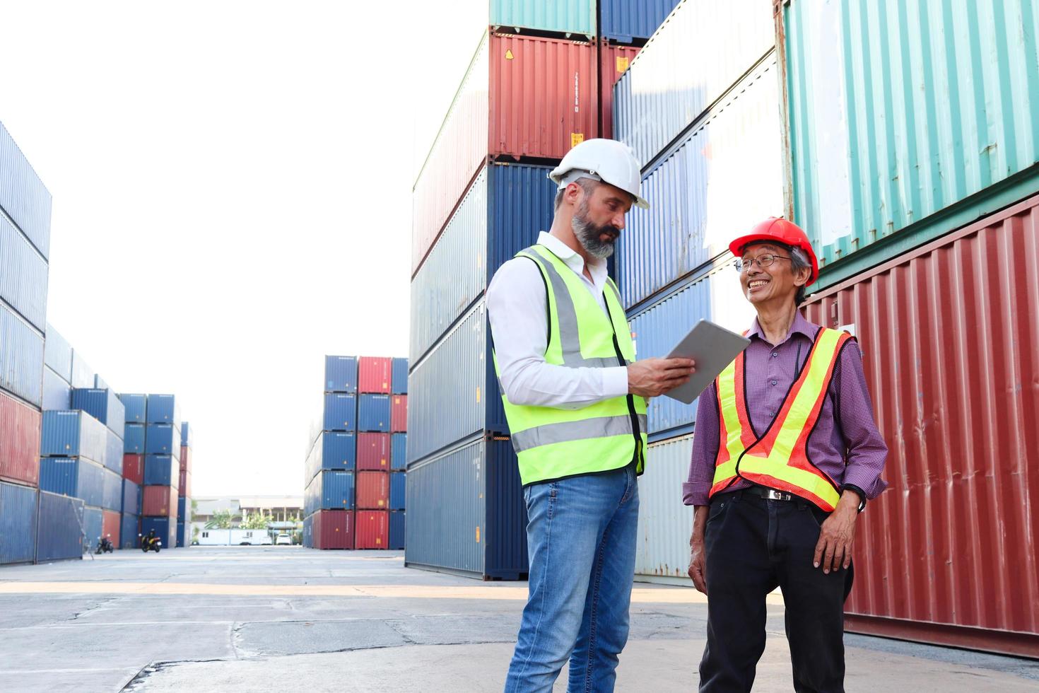 Two workers wearing safety vest and helmet discussing at logistic shipping cargo containers yard. serious senior elderly Asian worker engineer talking with his boss at workplace. photo