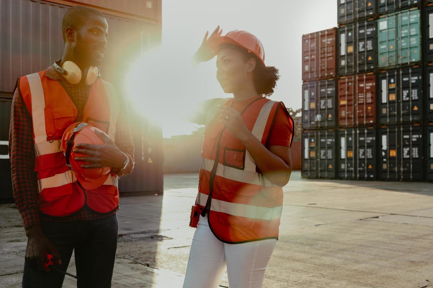 Happy smiling industrial African American engineer man and woman wear safety vest and helmet, two workers working at logistic shipping cargo container yard in twilight sunset time. photo