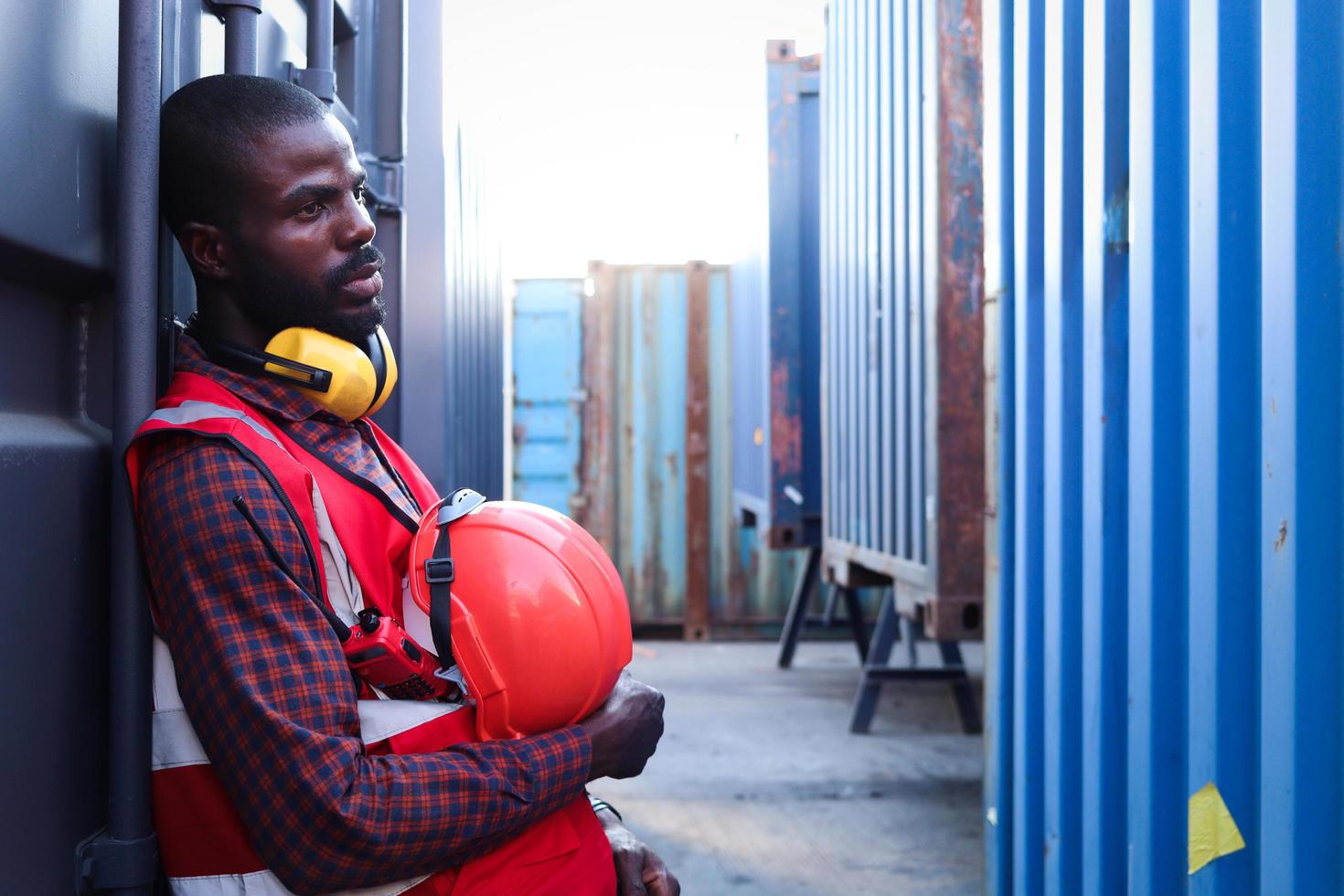 Portrait of African American young engineer worker man with safety red color vest and helmet, feeling tired from working hard, resting beside shipping container at logistic cargo yard. photo