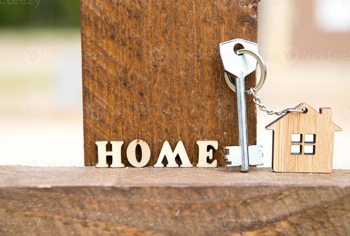 Keychain of wooden figure house on fence with key and inscription in English letters home. Cottage in the background. Building, project, moving to new house, mortgage, rent and purchase real estate. photo