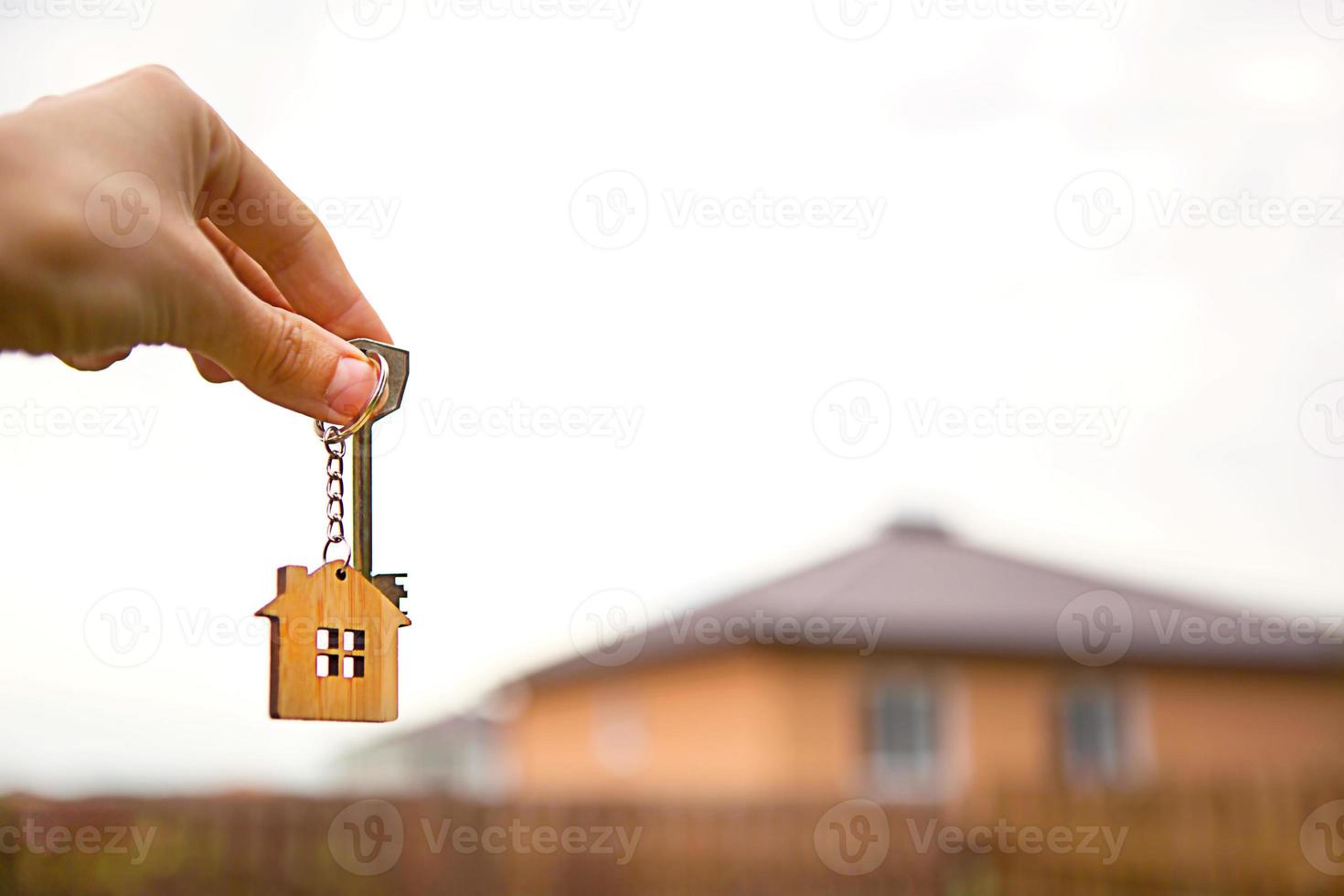 Hand with a key and a wooden key ring-house. Background of fence and cottage. Building, project, moving to a new home, mortgage, rent and purchase real estate. To open the door. Copy space photo