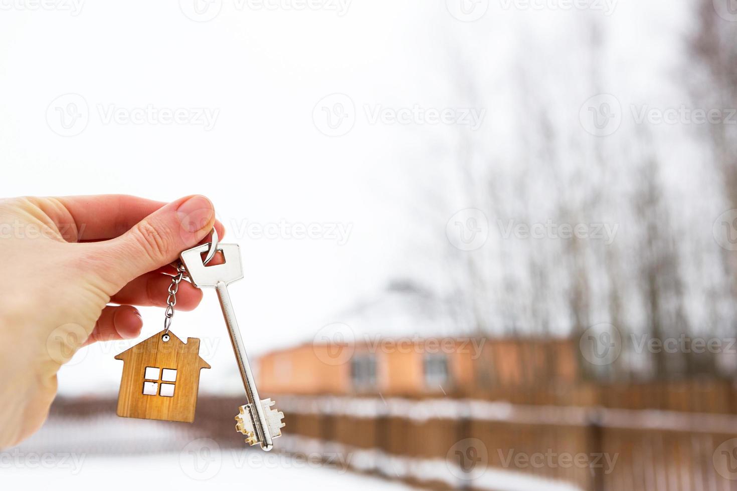 Hand with a Key of the house in winter in the snow. Background of fence and cottage. Give a gift for New Year, Christmas. Moving to new house, mortgage, rent and purchase real estate. Open the door. photo
