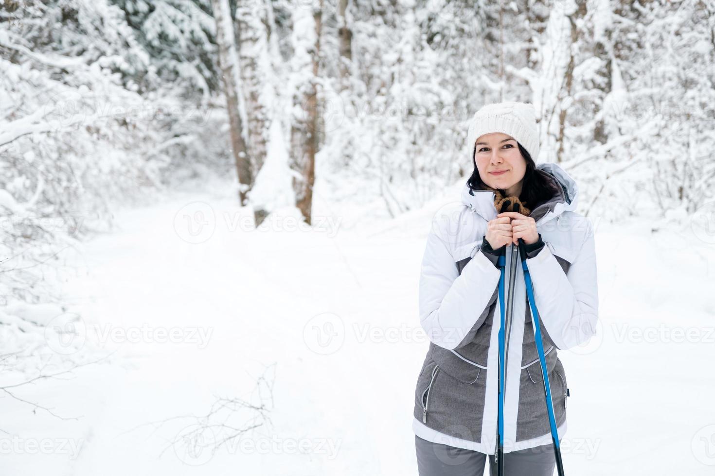 Skier a woman in a membrane jacket with ski poles in his hands with his back against the background of a snowy forest. Cross-country skiing in winter forest, outdoor sports, healthy lifestyle. photo