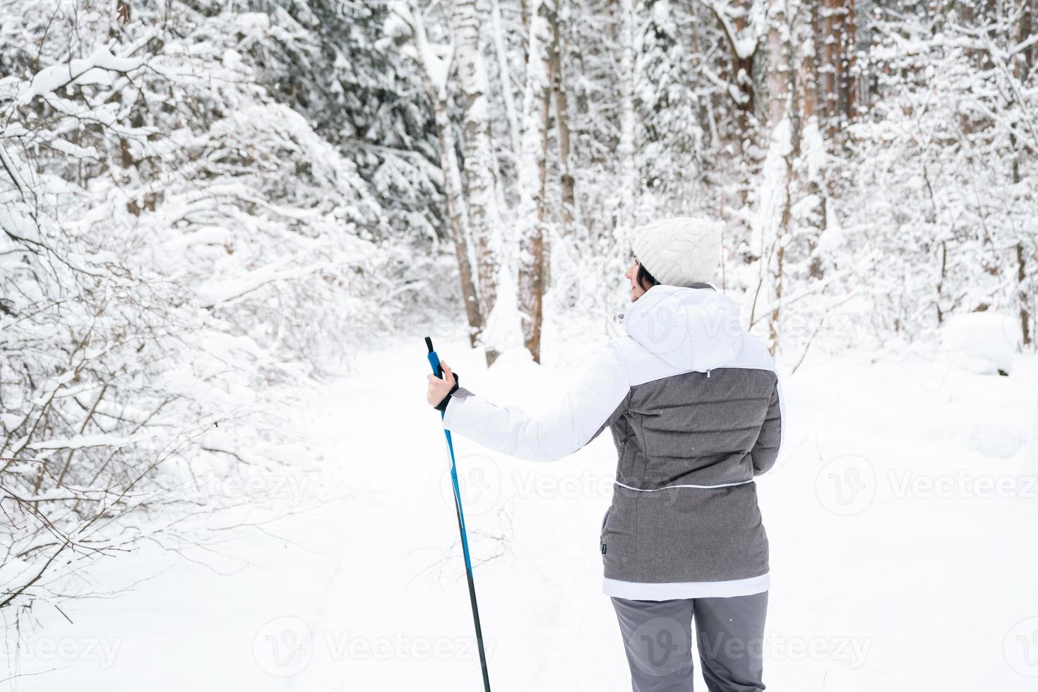 Skier a woman in a membrane jacket with ski poles in his hands with his back against the background of a snowy forest. Cross-country skiing in winter forest, outdoor sports, healthy lifestyle. photo