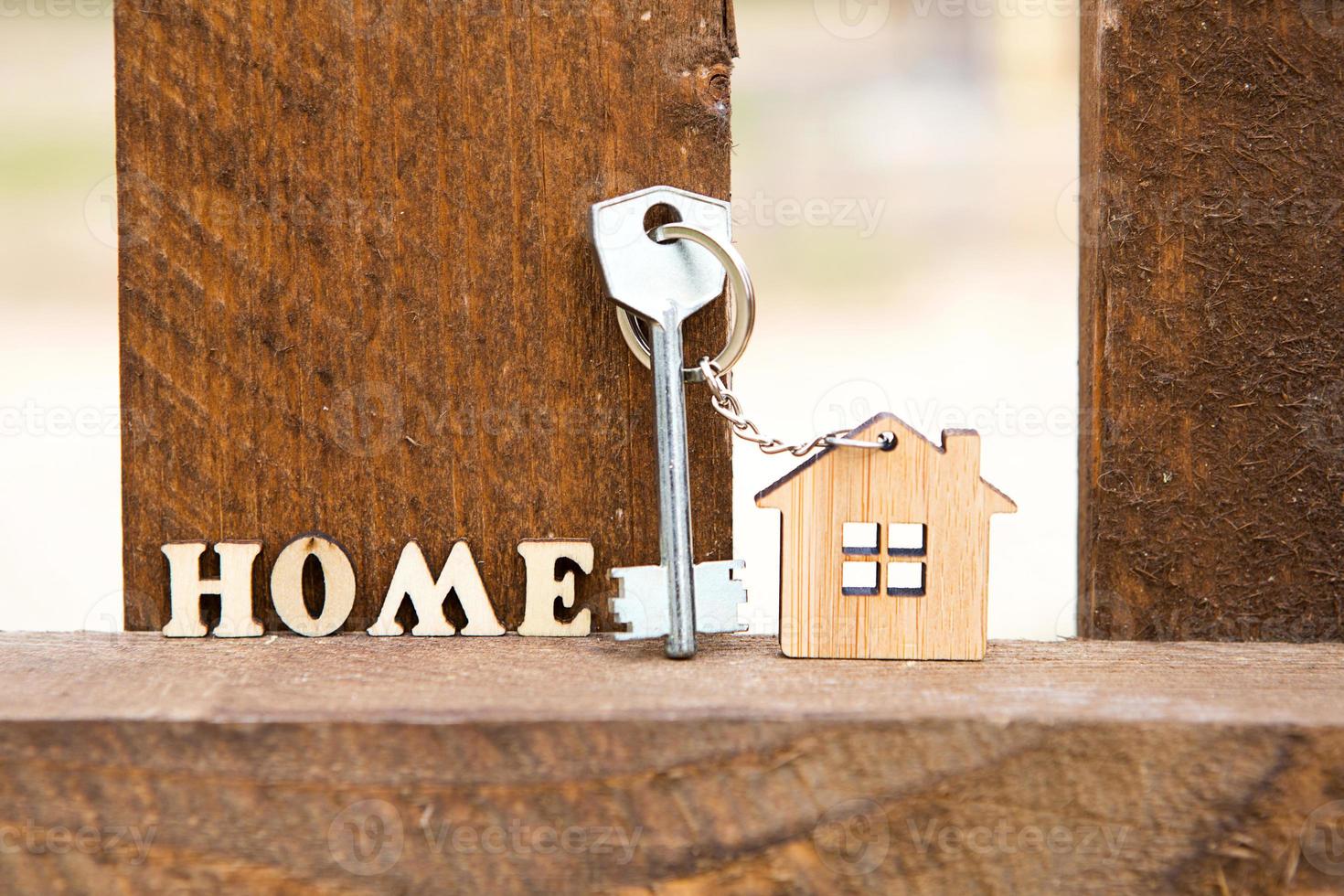 Keychain of wooden figure house on fence with key and inscription in English letters home. Cottage in the background. Building, project, moving to new house, mortgage, rent and purchase real estate. photo