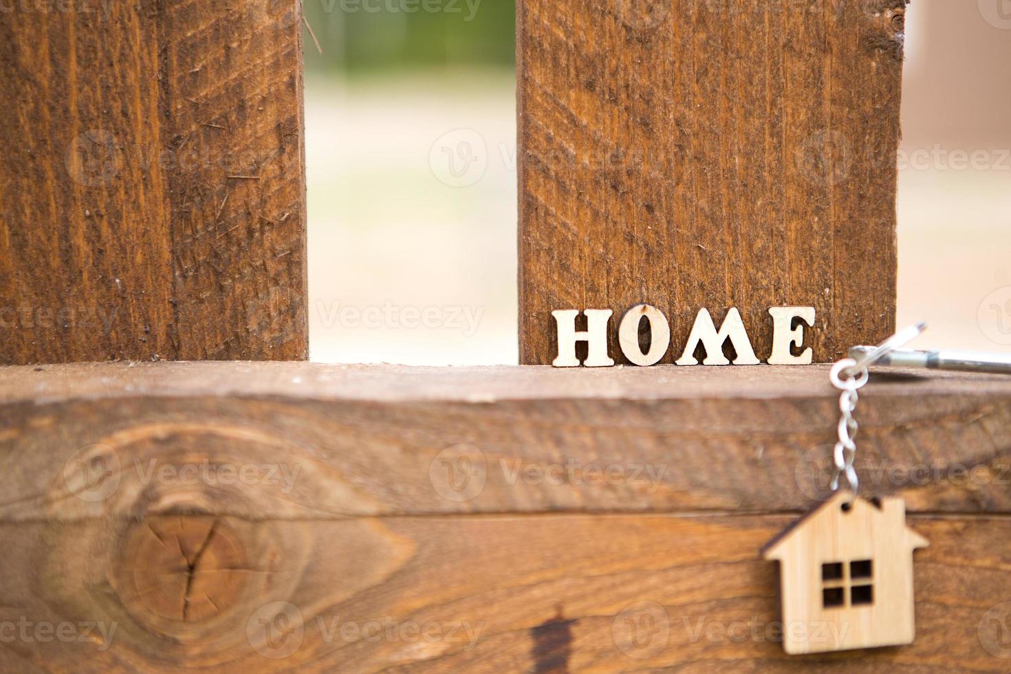 Keychain of wooden figure house on fence with key and inscription in English letters home. Cottage in the background. Building, project, moving to new house, mortgage, rent and purchase real estate. photo