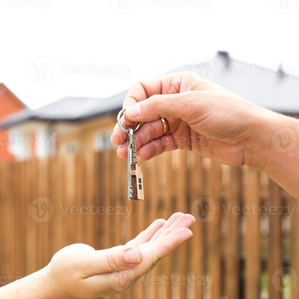 Hand with a key and a wooden key ring-house. Background of fence and cottage. Building, project, moving to a new home, mortgage, rent and purchase real estate. To open the door. Copy space photo