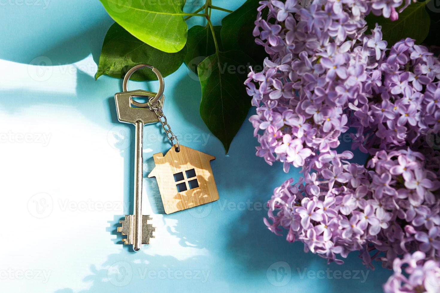 House key with keychain on blue spring background and lilac branches.Summer holiday home, booking cottage in the countryside, moving to new home, mortgage, rent and purchase real estate. Tourism photo