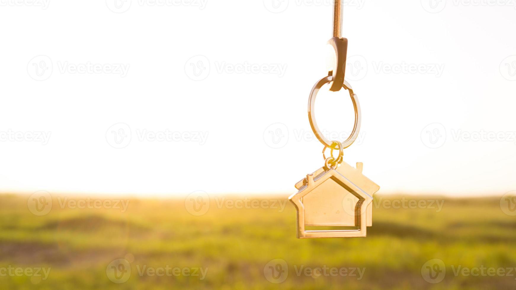 House key with keychain in hand. Background of sky, sunlight and field. Dream of home, building a cottage in countryside, plan and project design, farm, moving to a new home, accommodation. Copy space photo