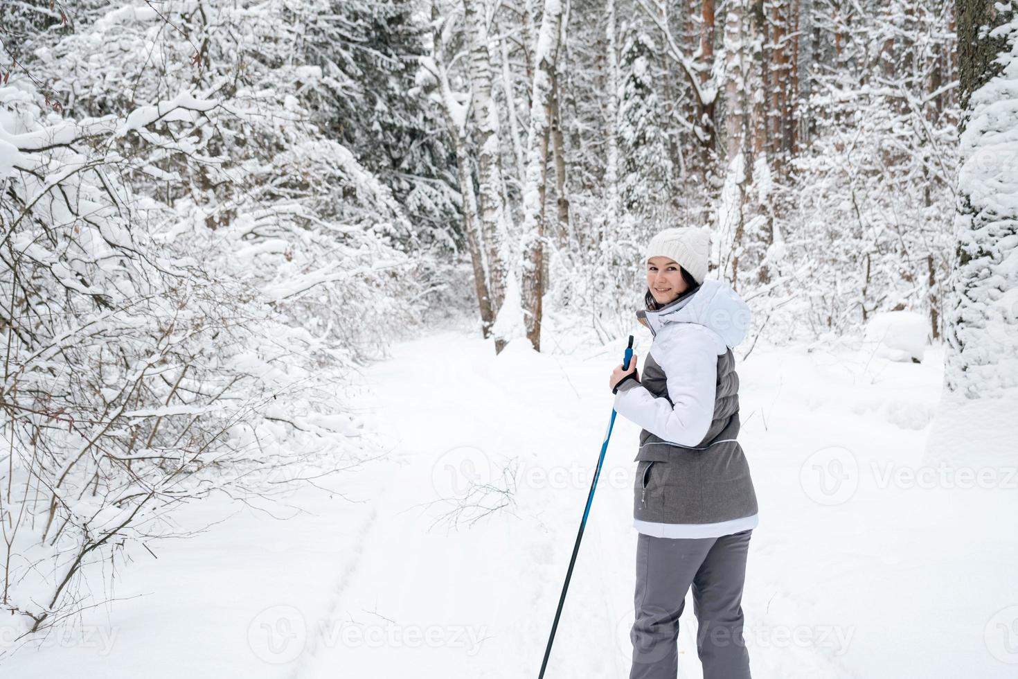 Skier a woman in a membrane jacket with ski poles in his hands with his back against the background of a snowy forest. Cross-country skiing in winter forest, outdoor sports, healthy lifestyle. photo