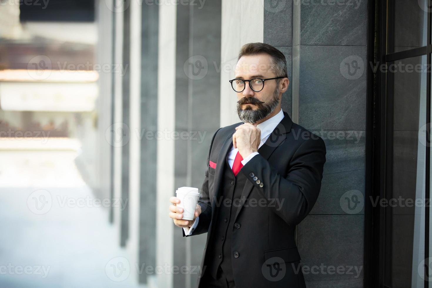 hombre de negocios hipster en traje de pie con una taza de café cerca de la oficina al aire libre foto