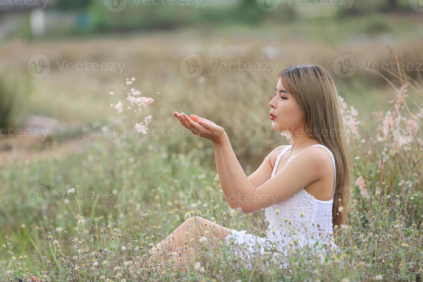 hermosa mujer joven sentada en el campo de hierba verde y soplando diente de león. al aire libre. disfruta de la naturaleza. niña sonriente saludable en el césped de primavera. concepto libre de alergias. libertad foto