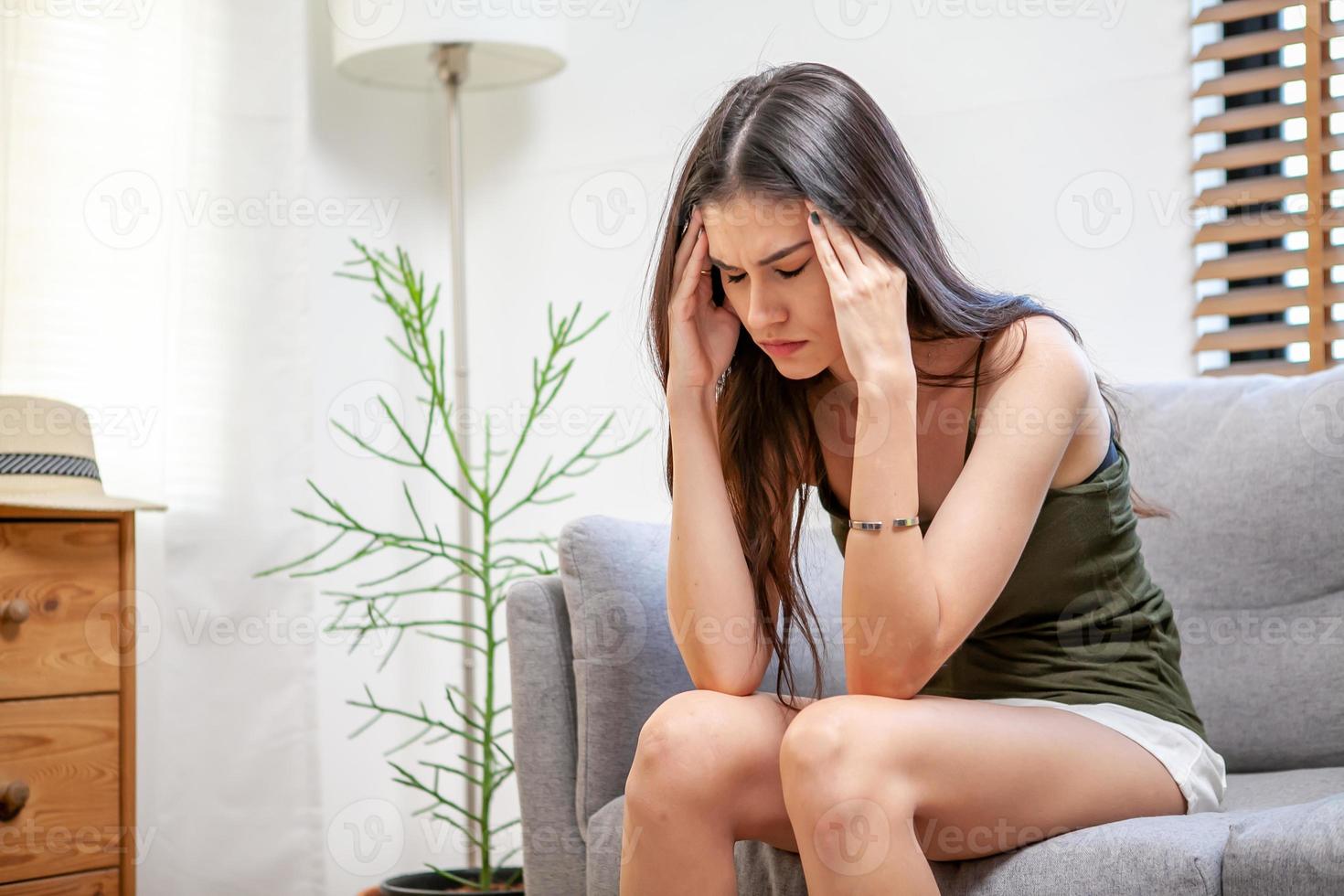 triste mujer joven deprimida en casa sentada en el sofá. niña frustrada e infeliz con terrible migraña o dolor de cabeza. foto
